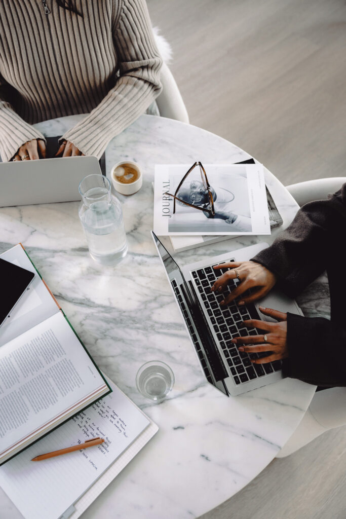 Two women working on laptops at a marble table with journals, coffee, and handwritten notes—creative business planning workspace