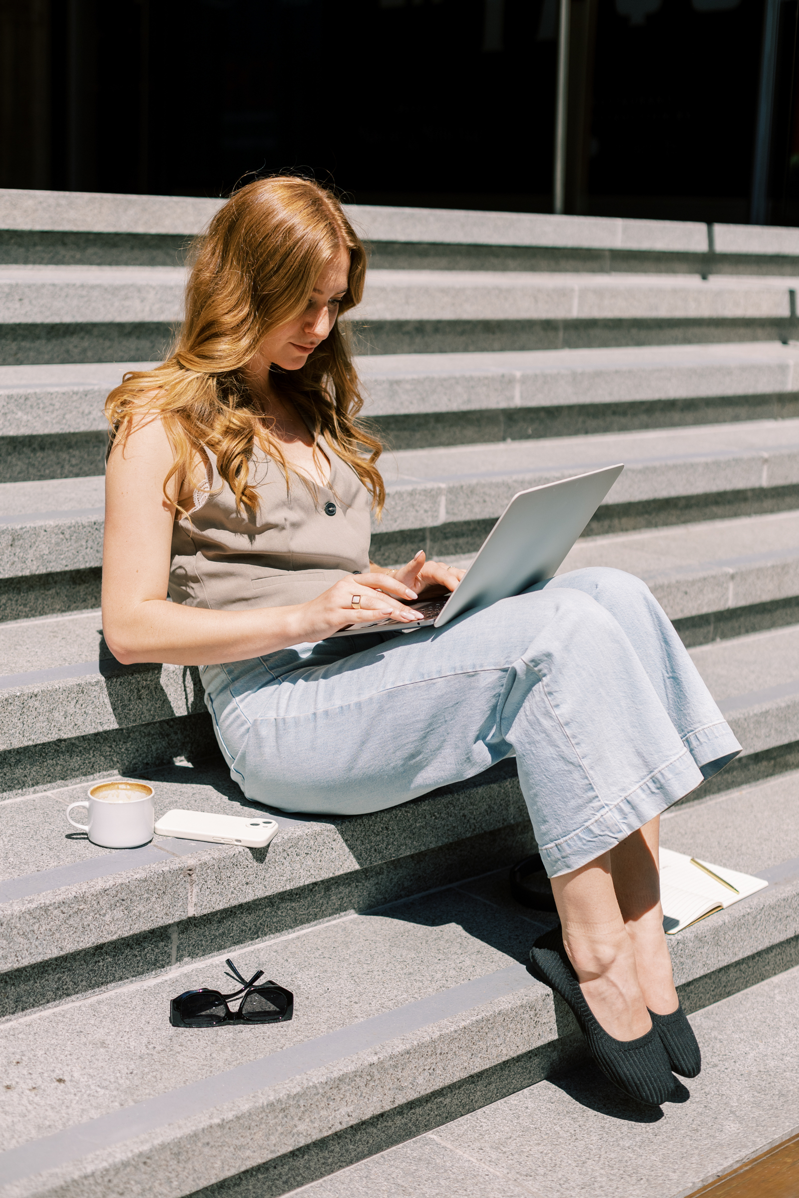Woman entrepreneur working on laptop outdoors with coffee, phone, and notebook beside her.
