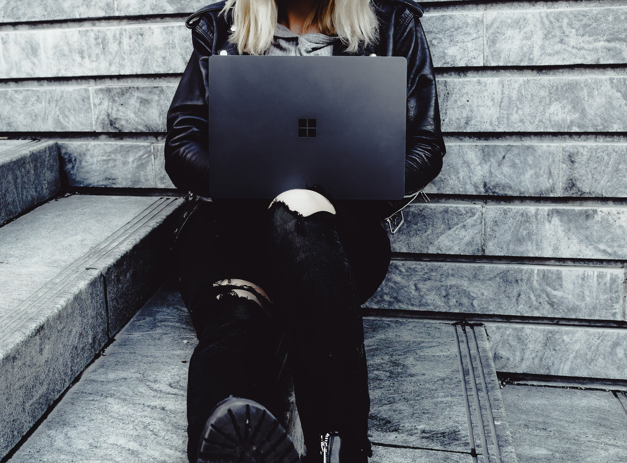 Young woman sitting on outdoor stone steps working on a Microsoft Surface laptop, dressed in black with ripped jeans and leather jacket