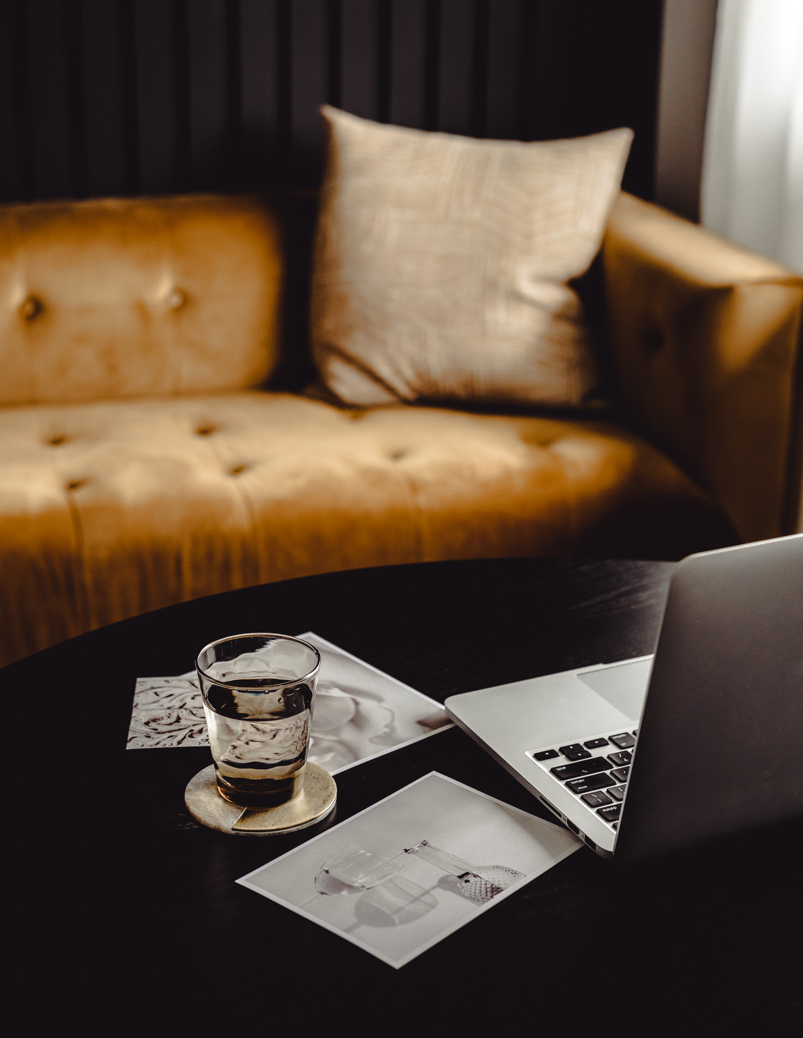 Modern workspace with a laptop, water glass, and art prints on a black round table beside a gold velvet couch.