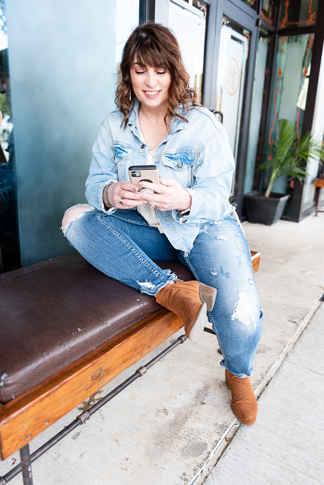 Jan Touchberry sitting on an outdoor bench, smiling while using her phone, wearing a light denim jacket and brown ankle boots
