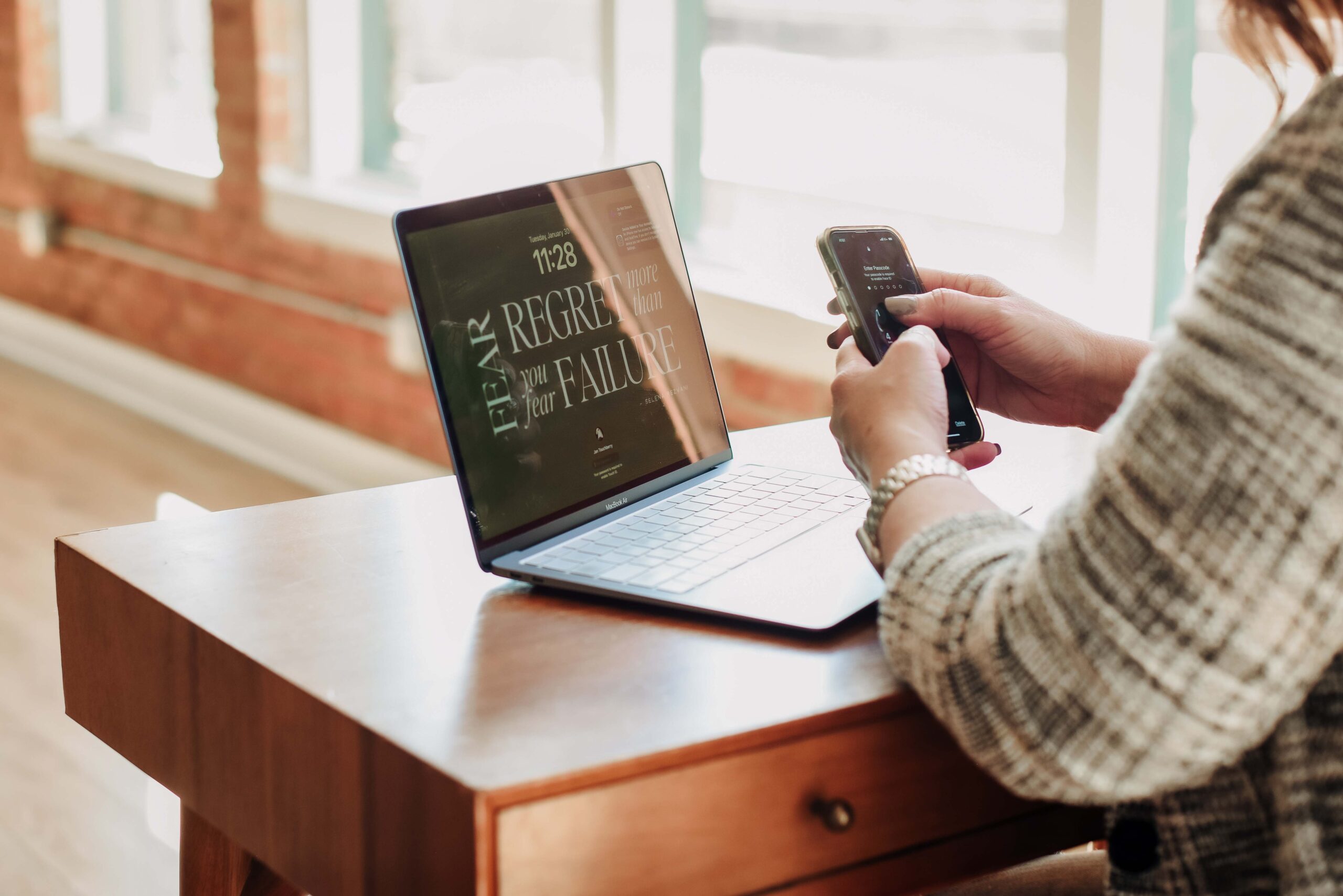 Woman seated at a desk by a bright window, working on a laptop with a notebook and pen beside her, surrounded by plants.