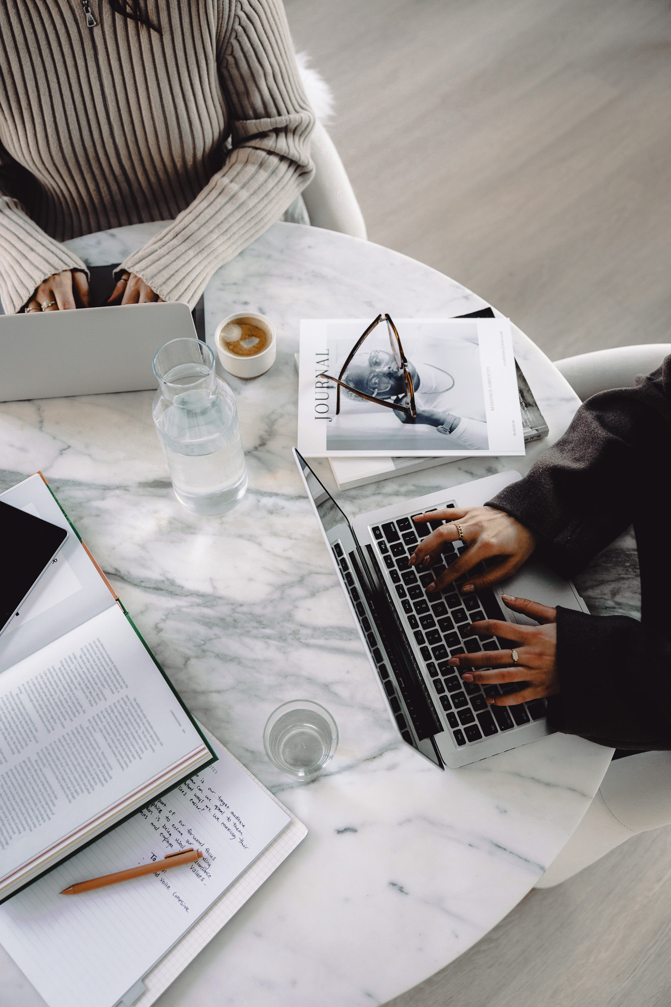 Two women working on laptops at a marble table with journals, coffee, and handwritten notes—creative business planning workspace